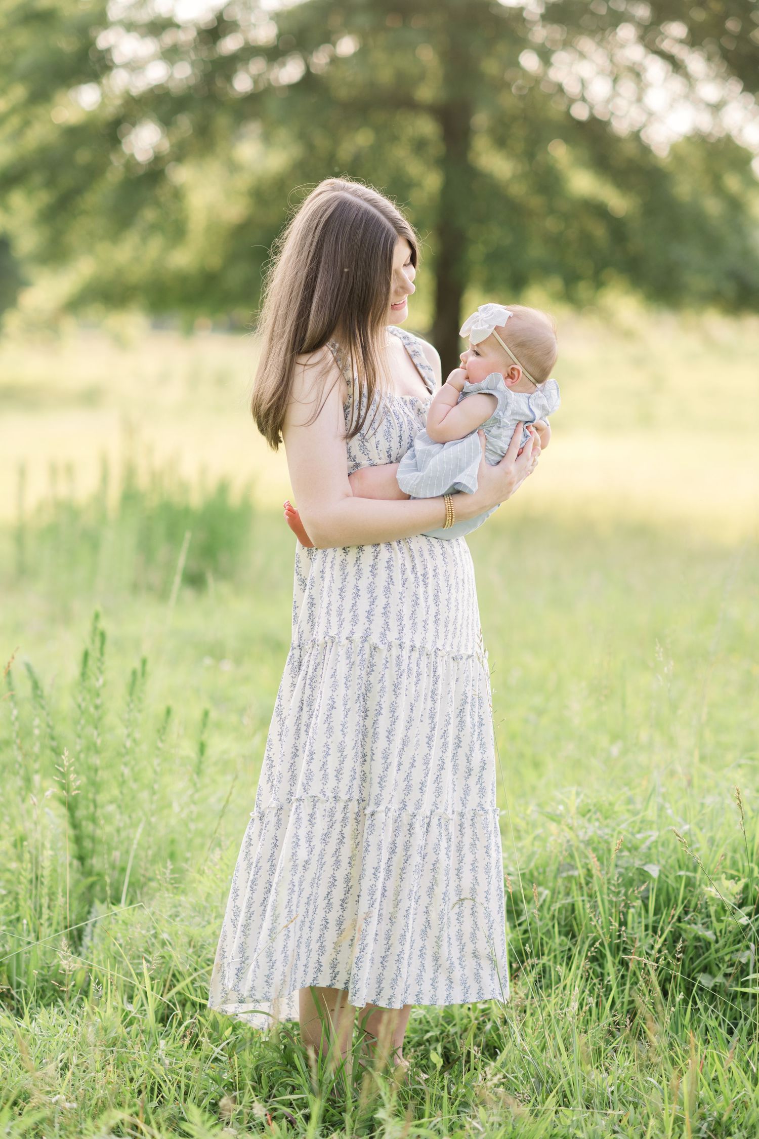 A mother in a floral midi dress holds her infant baby while standing in a lush green meadow.