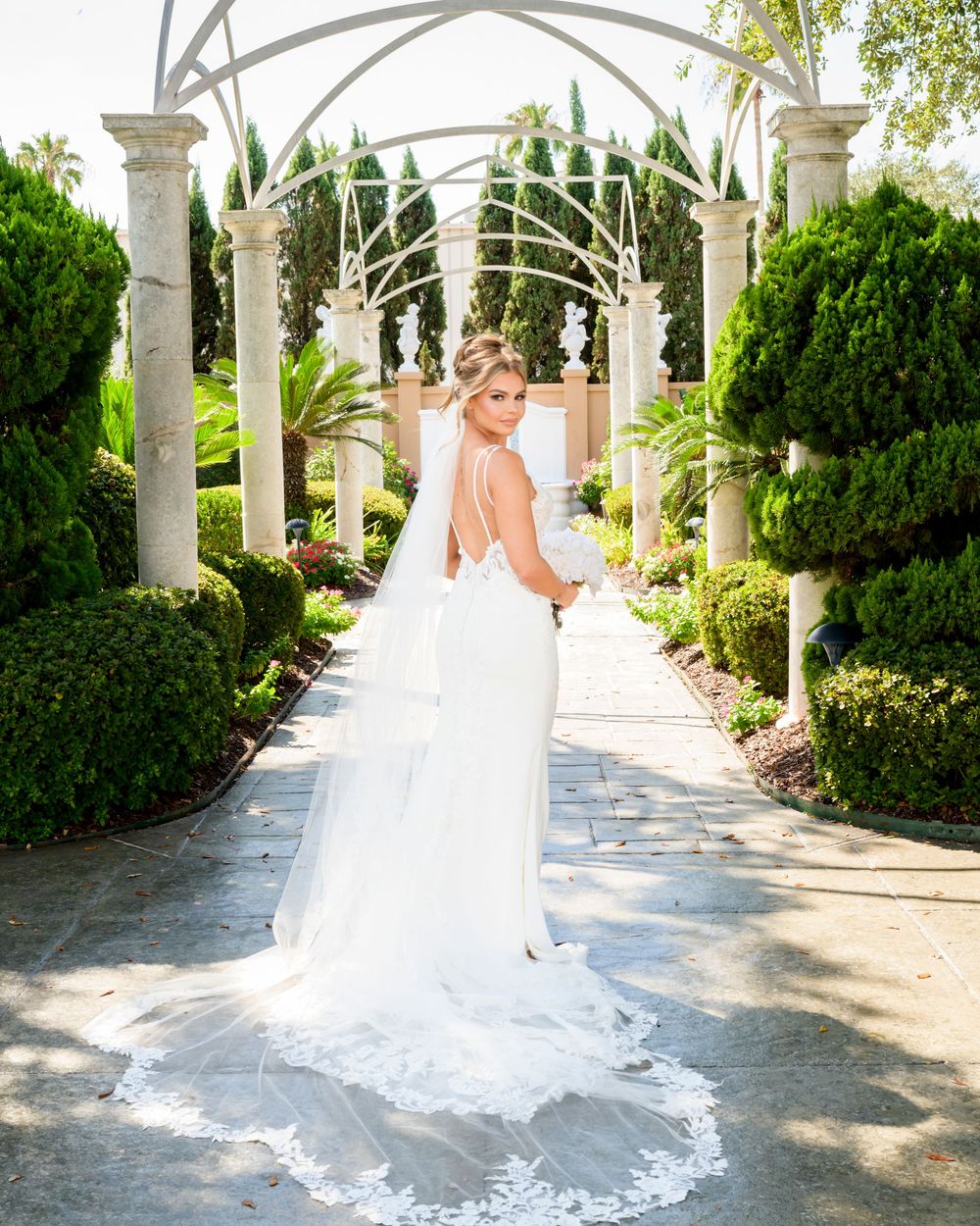 Beautiful bride in a flowing white wedding gown poses gracefully under an elegant garden pergola with manicured landscaping.