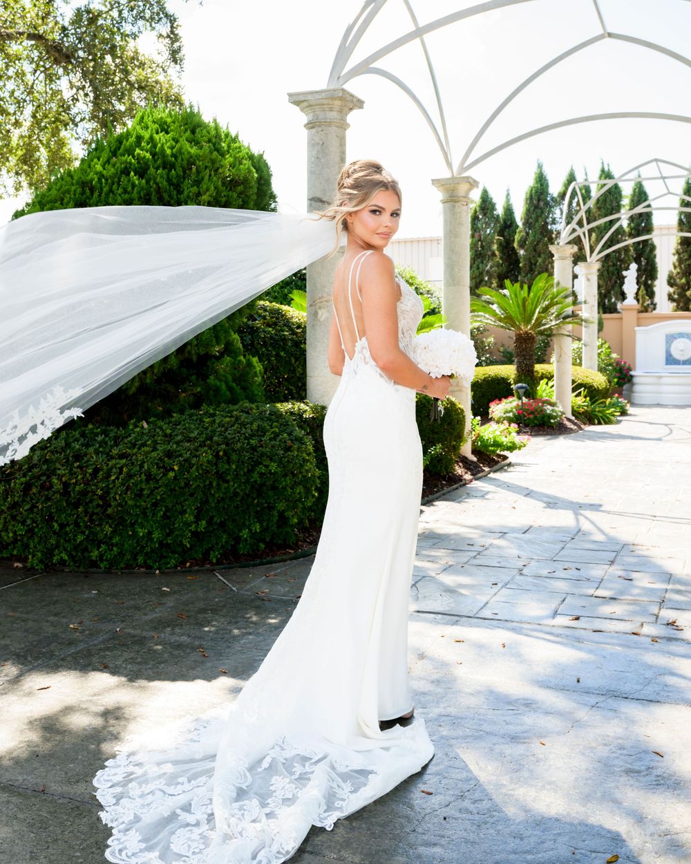 Elegant white wedding dress with flowing veil and train photographed in a beautiful garden setting with white columns.