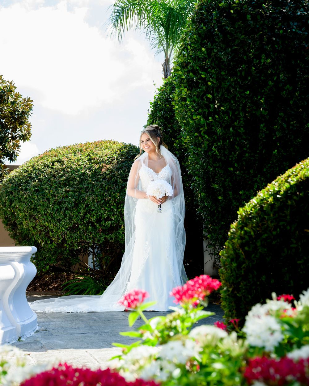 Elegant bride poses in white wedding gown and veil against lush garden backdrop with colorful flowers.