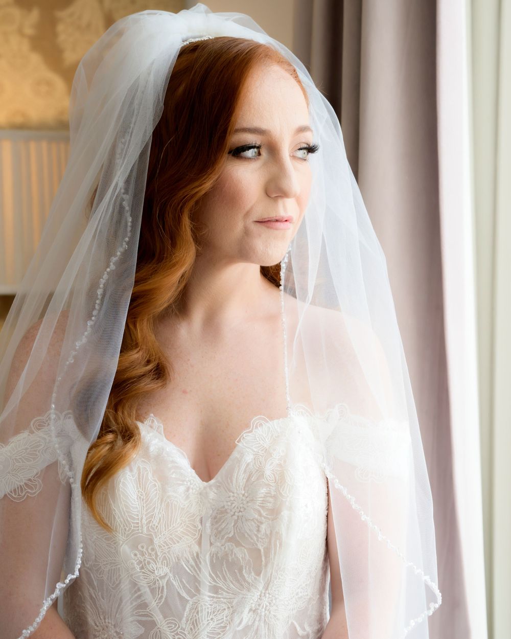 A bride in a white lace wedding dress and long veil poses near a window with natural lighting.