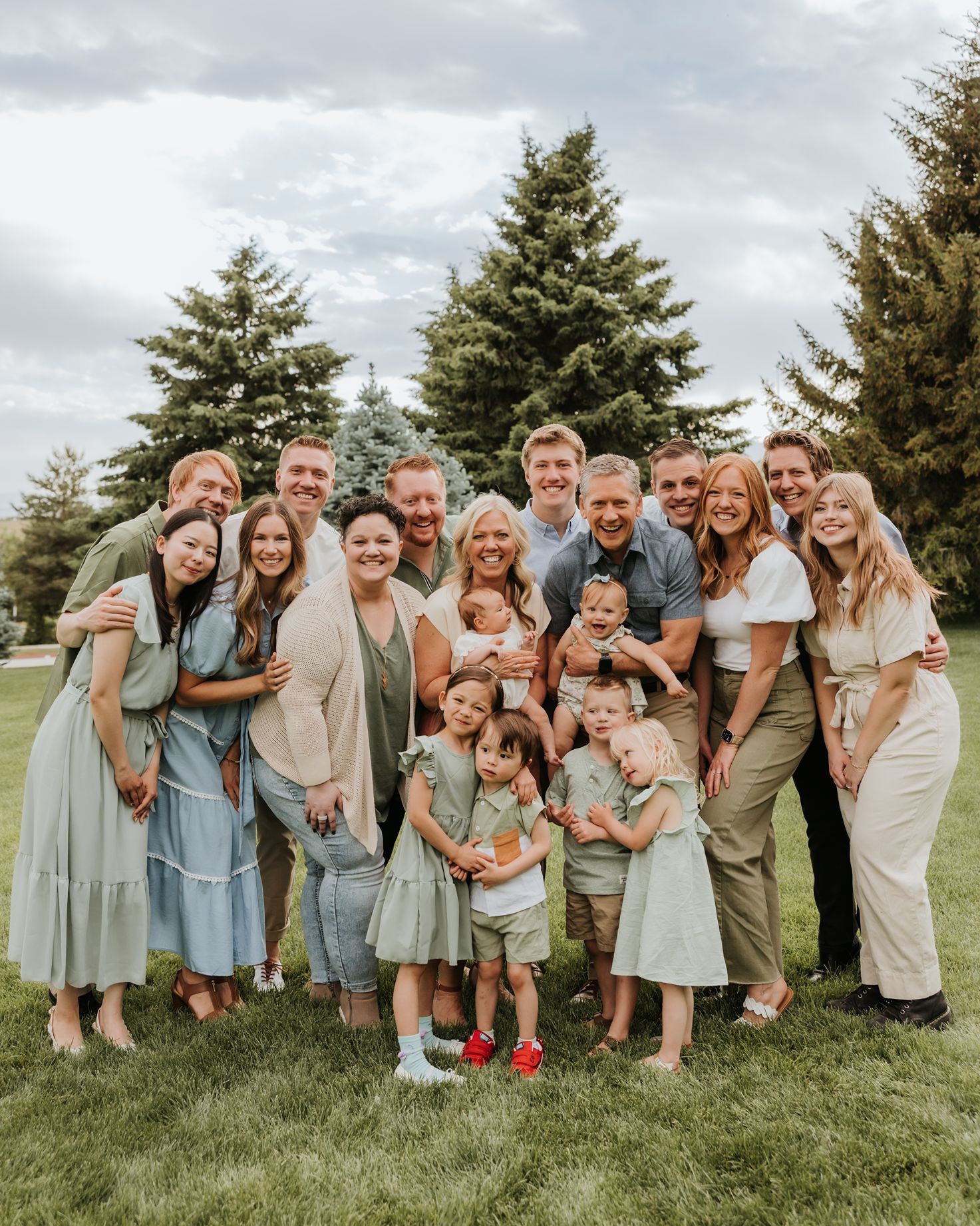 Utah Large extended family group poses playfully in coordinated blue and beige outfits on grassy lawn with evergreen trees.