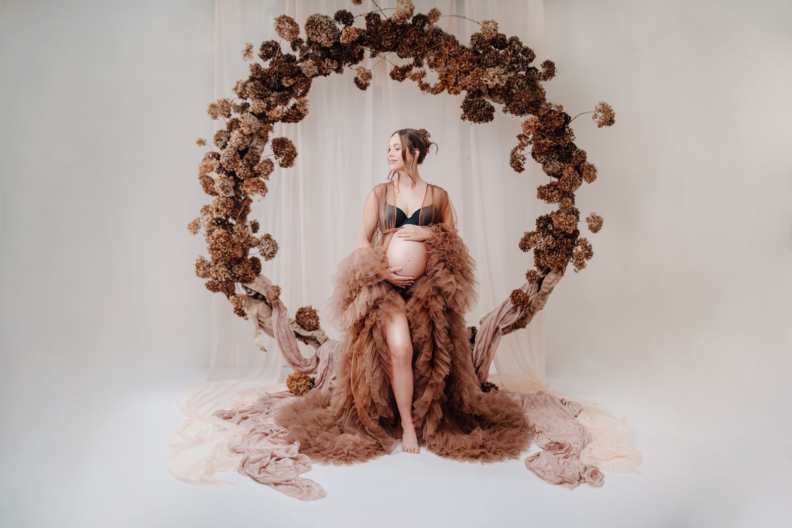 A romantic maternity photo featuring a floral arch backdrop and flowing dusty rose fabric draping  Babybauch Fotografie Hessen
