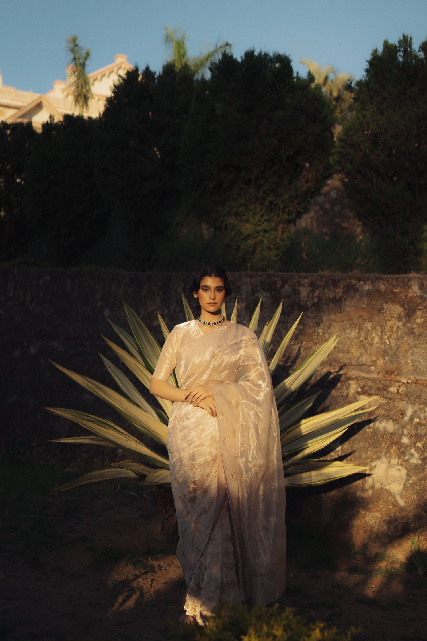 Figure in elegant white sari standing against agave plant at dusk.
