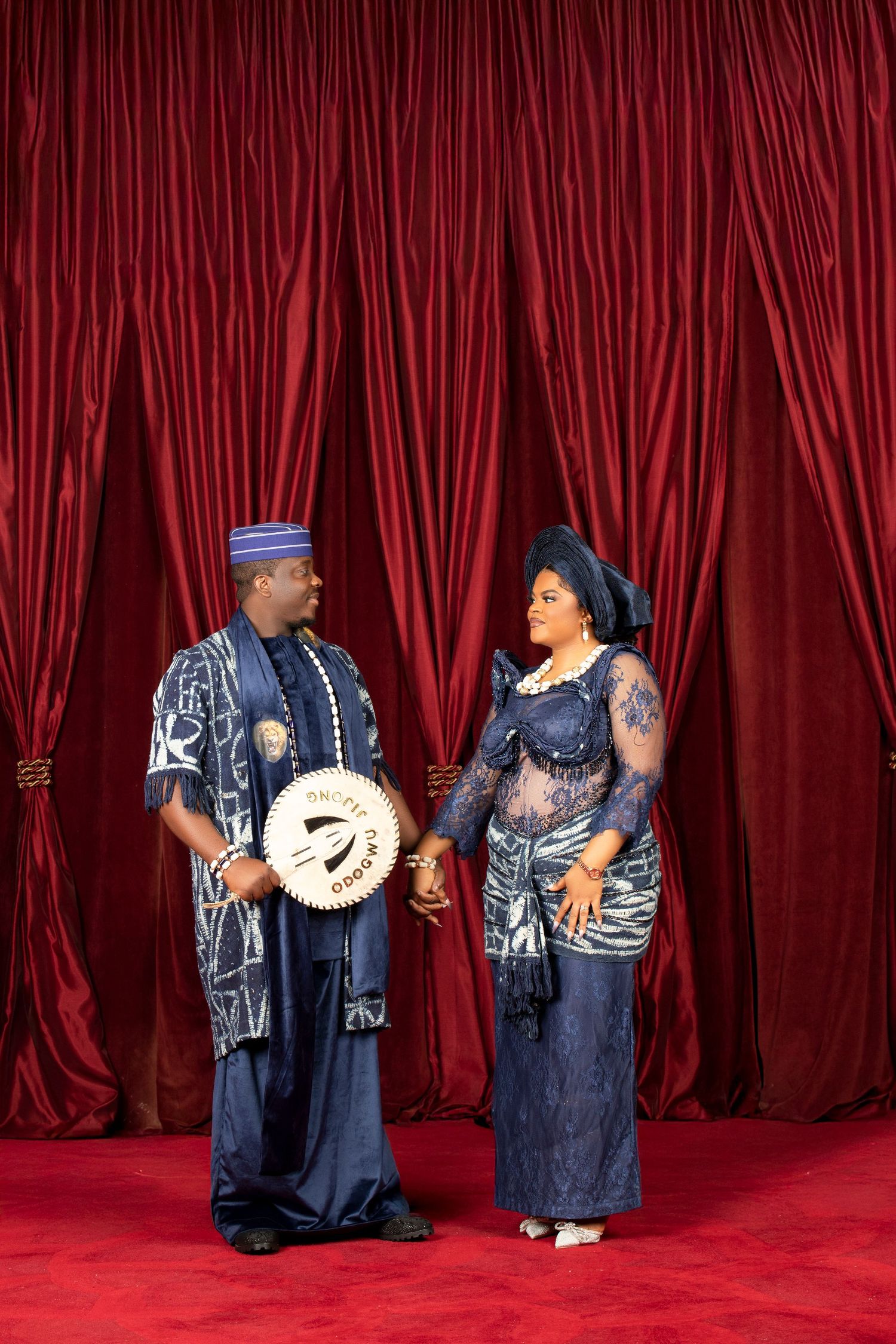Traditional Nigerian formal attire portrait with couple in blue outfits against red curtain backdrop.