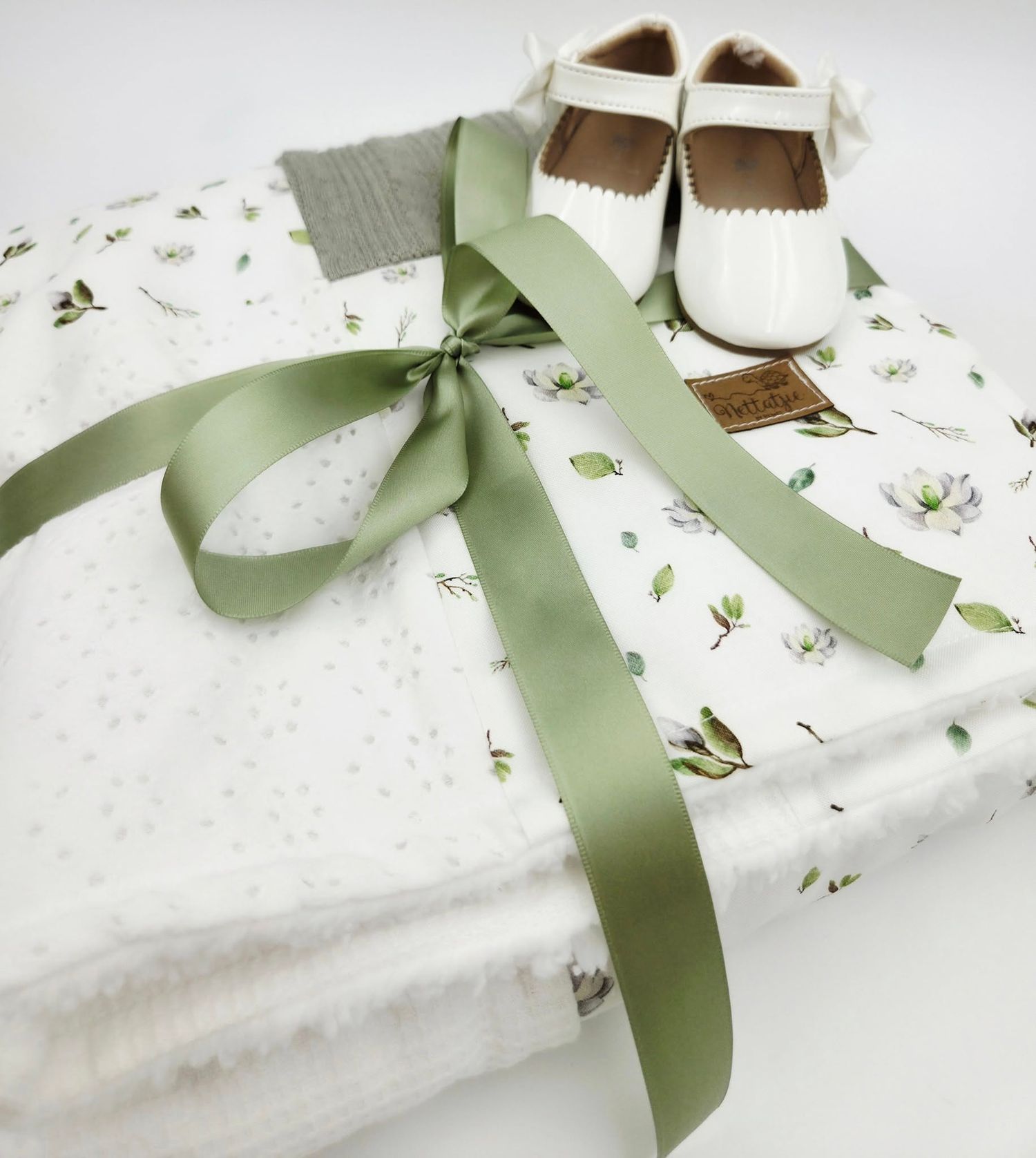 White floral baby blanket with sage green ribbon and tiny white leather shoes.