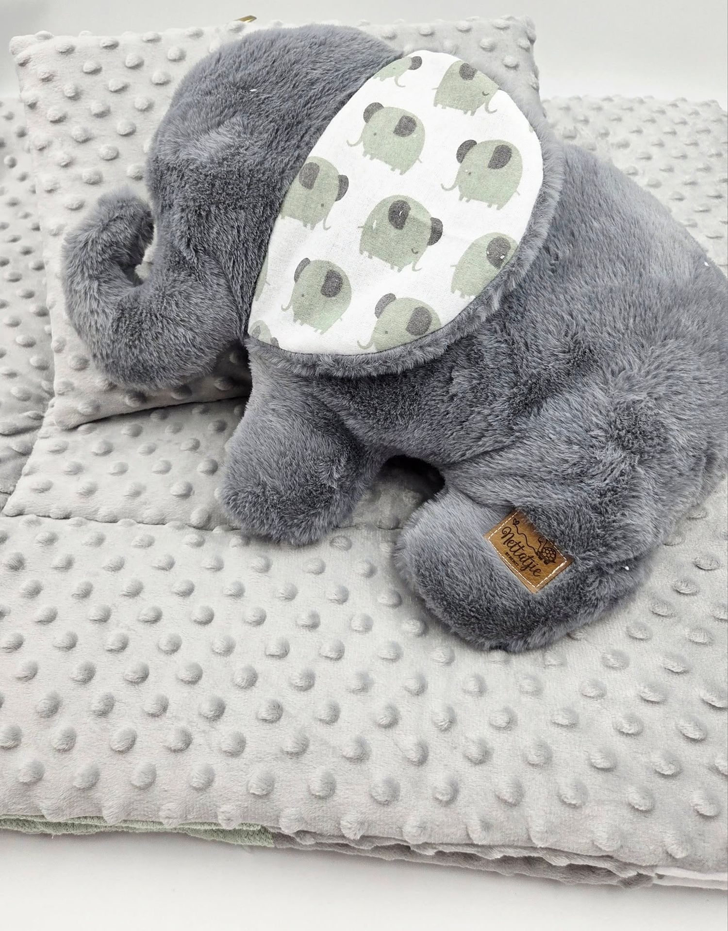 Plush gray elephant toy with patterned ears sitting on white dotted blanket.