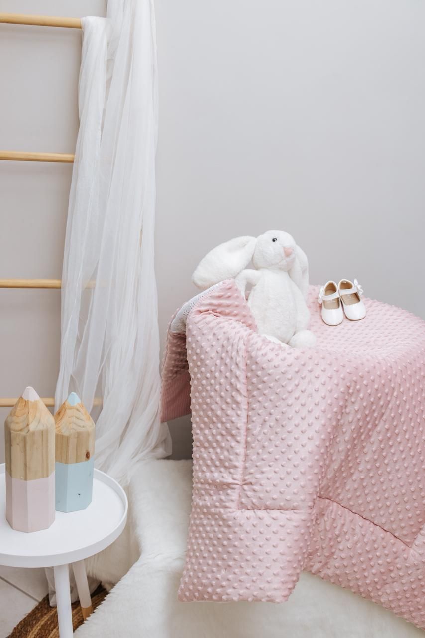 White and pink nursery decor with stuffed bunny, baby shoes and textured blanket.