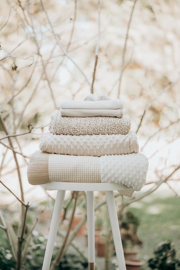 Stack of neutral beige and cream folded blankets and towels on a white stool outdoors in a garden.