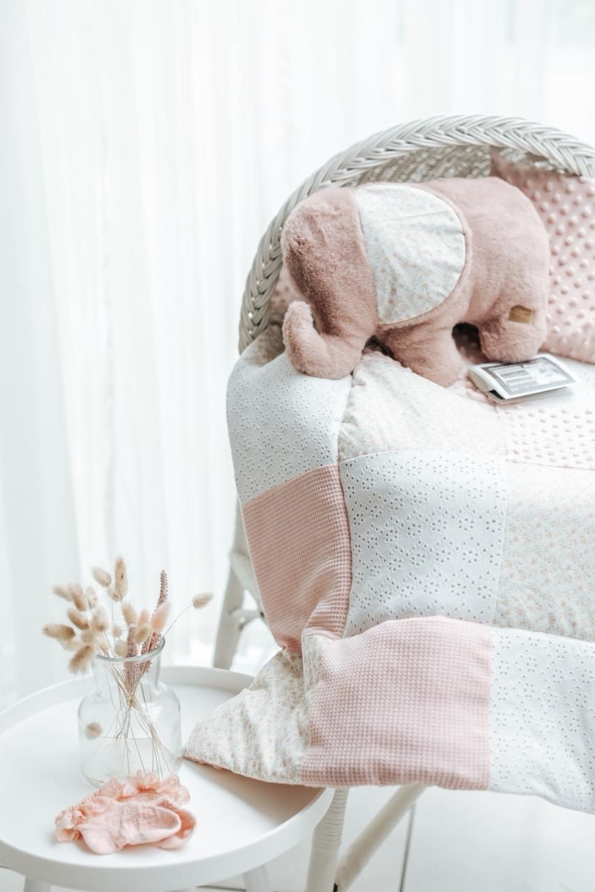 Soft pink and white nursery decor with plush elephant toy and textured pillows on a chair beside delicate dried flowers.