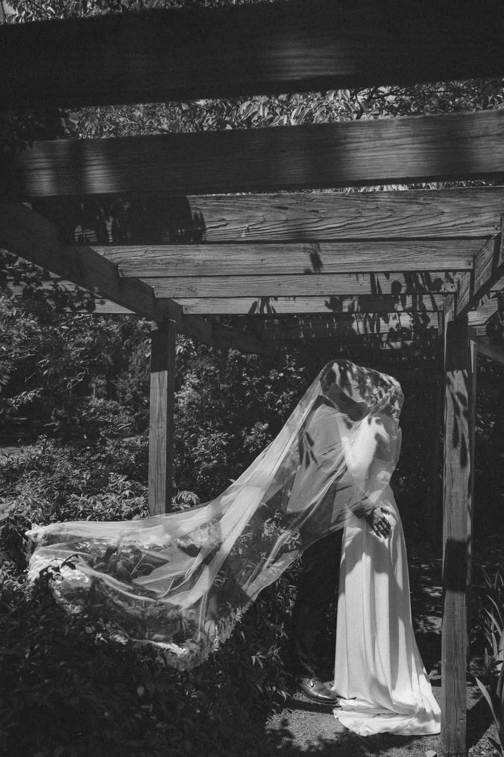 Black and white photo of a bride in a flowing wedding dress and veil standing under a wooden pergola outdoors.