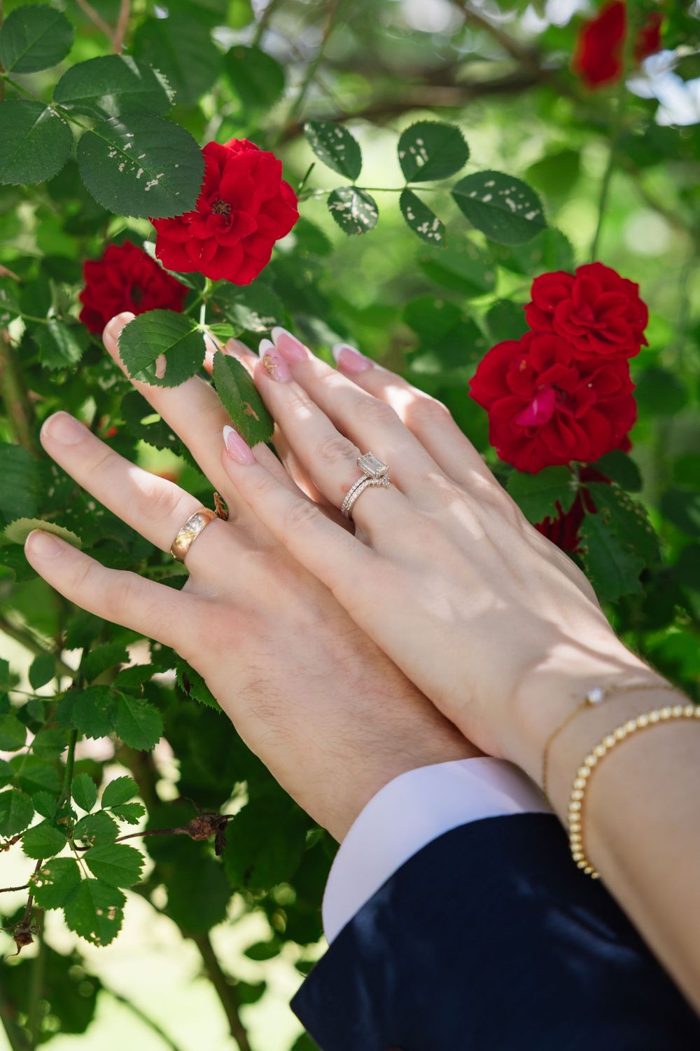 Newlywed couple displaying wedding rings and bracelet against vibrant red roses and green leaves in sunlit garden.
