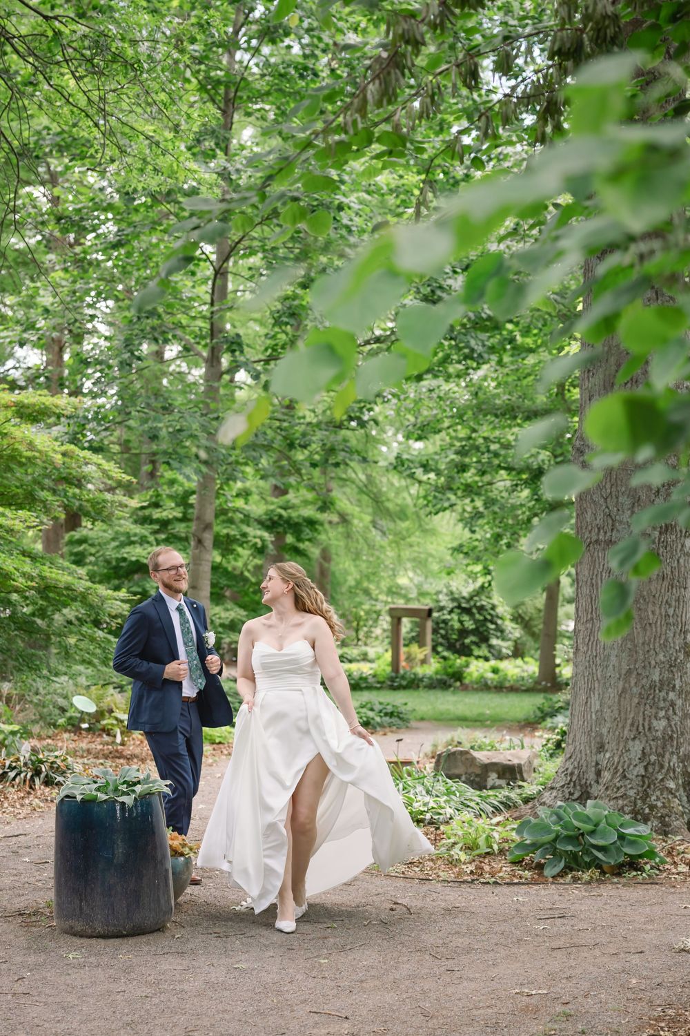 Bride and groom walk hand in hand through a lush green garden, bride's white gown flowing in the breeze.