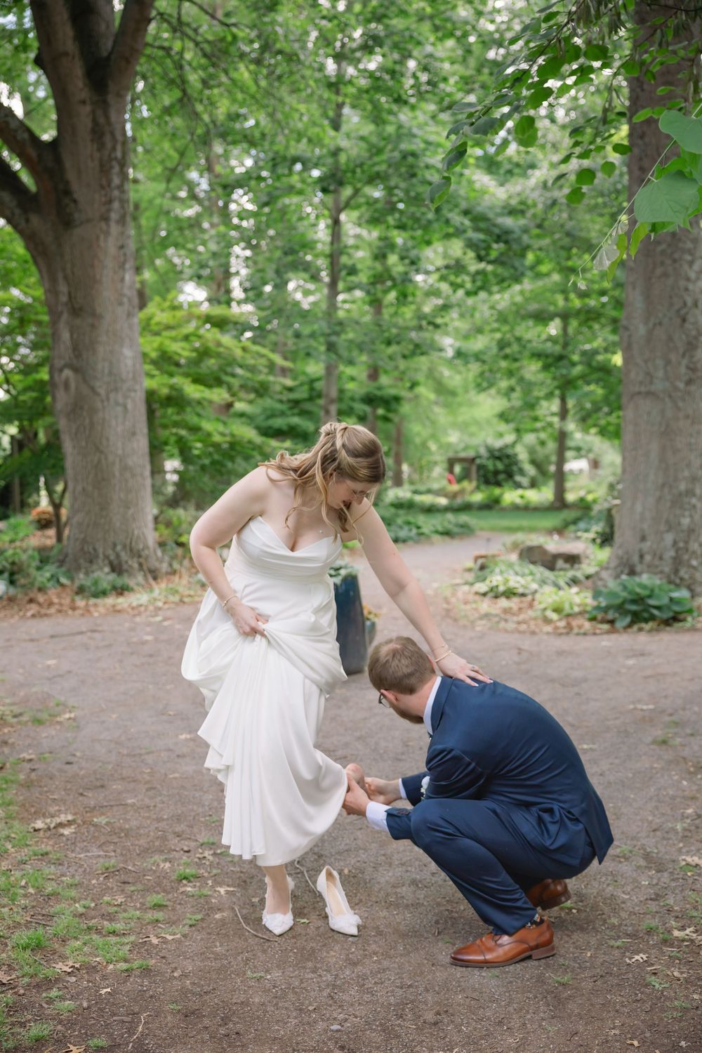Groom in navy suit fixes bride's white heel shoe in lush green park setting during outdoor wedding photo session.