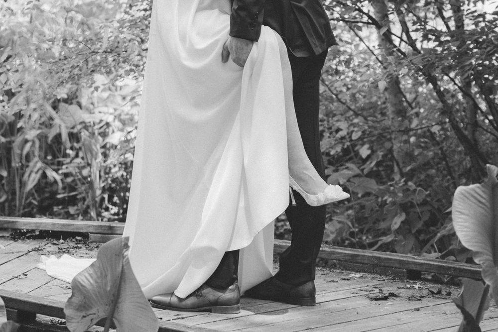 Black and white photo of a bride in a flowing white gown and groom in dark attire standing on a wooden deck outdoors.