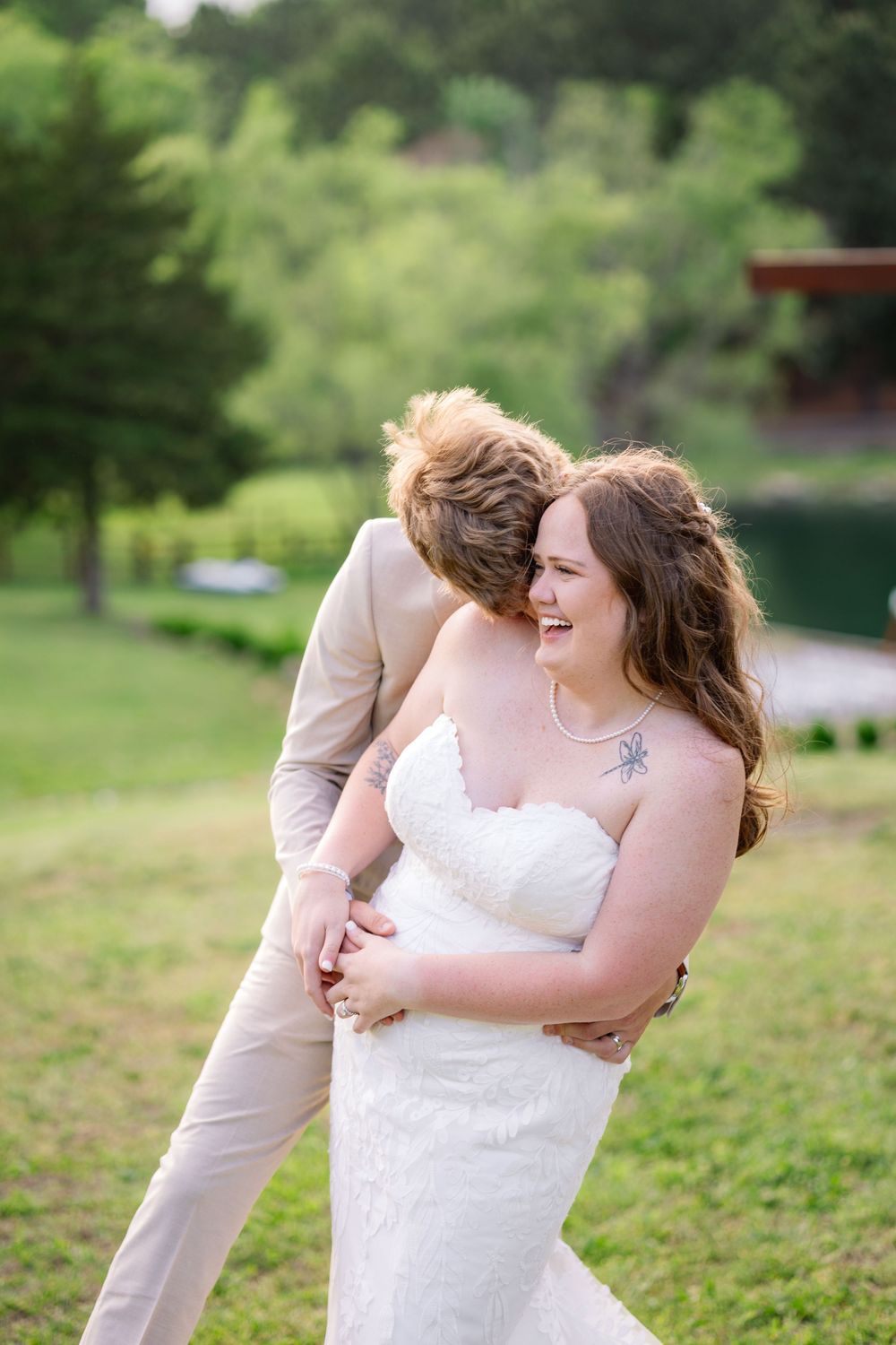 Bride in white strapless gown laughs as partner in tan suit embraces her from behind outdoors.