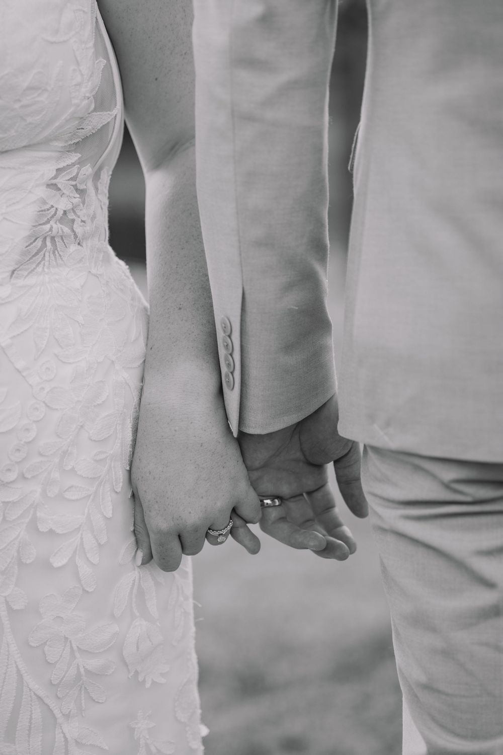 Black and white photo of newlyweds holding hands, showing wedding rings, bride in lace gown and groom in suit.