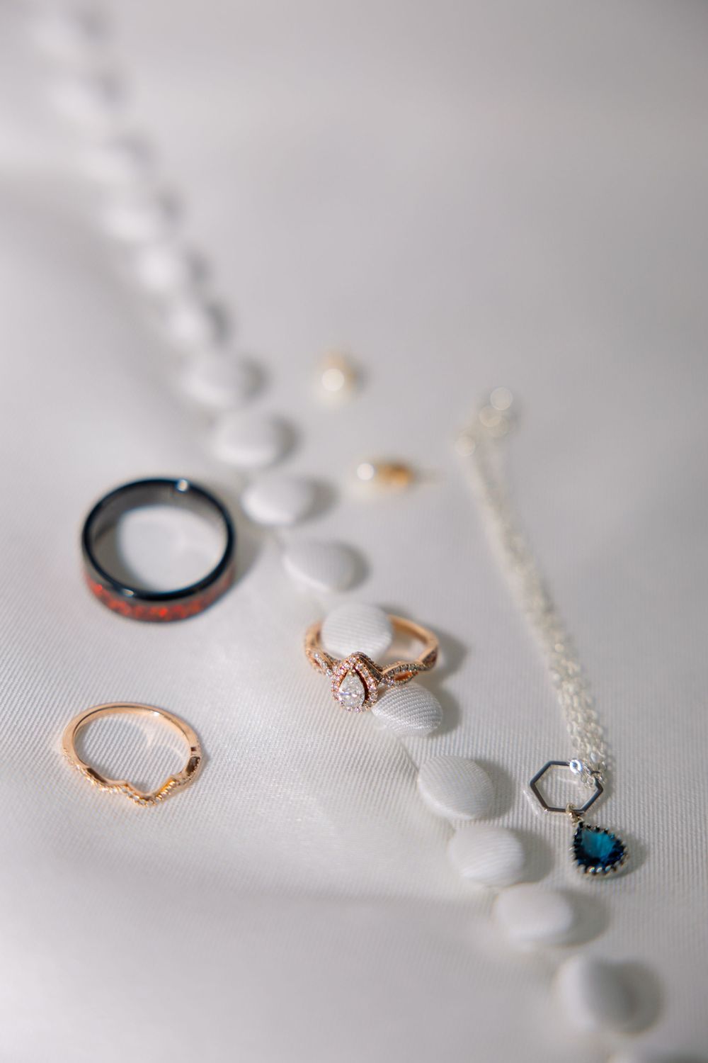 Elegant wedding jewelry flat lay featuring gold rings, a diamond engagement ring, a blue pendant necklace, and white beads on white fabric.