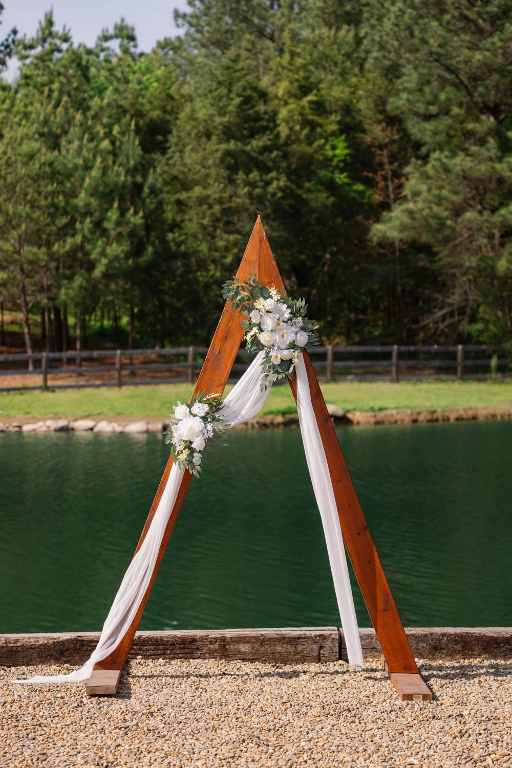 Rustic wooden triangular wedding arch adorned with white flowers and draped fabric beside a serene green lake.