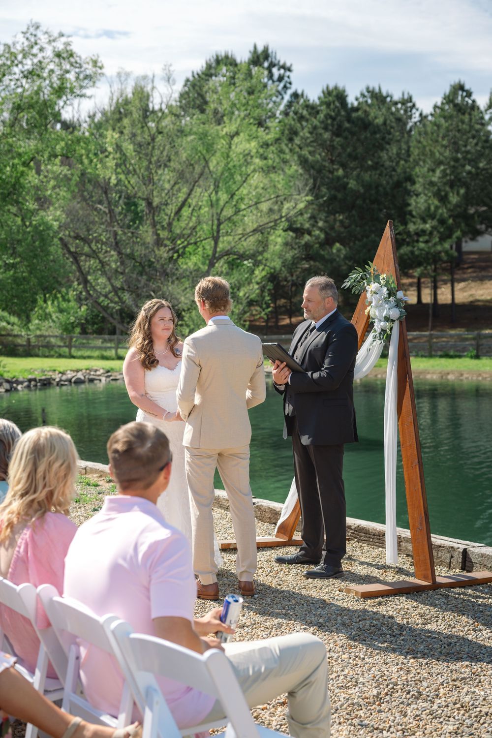 Outdoor wedding ceremony by a lake with bride, groom, and officiant at a wooden arch, guests seated in white chairs.