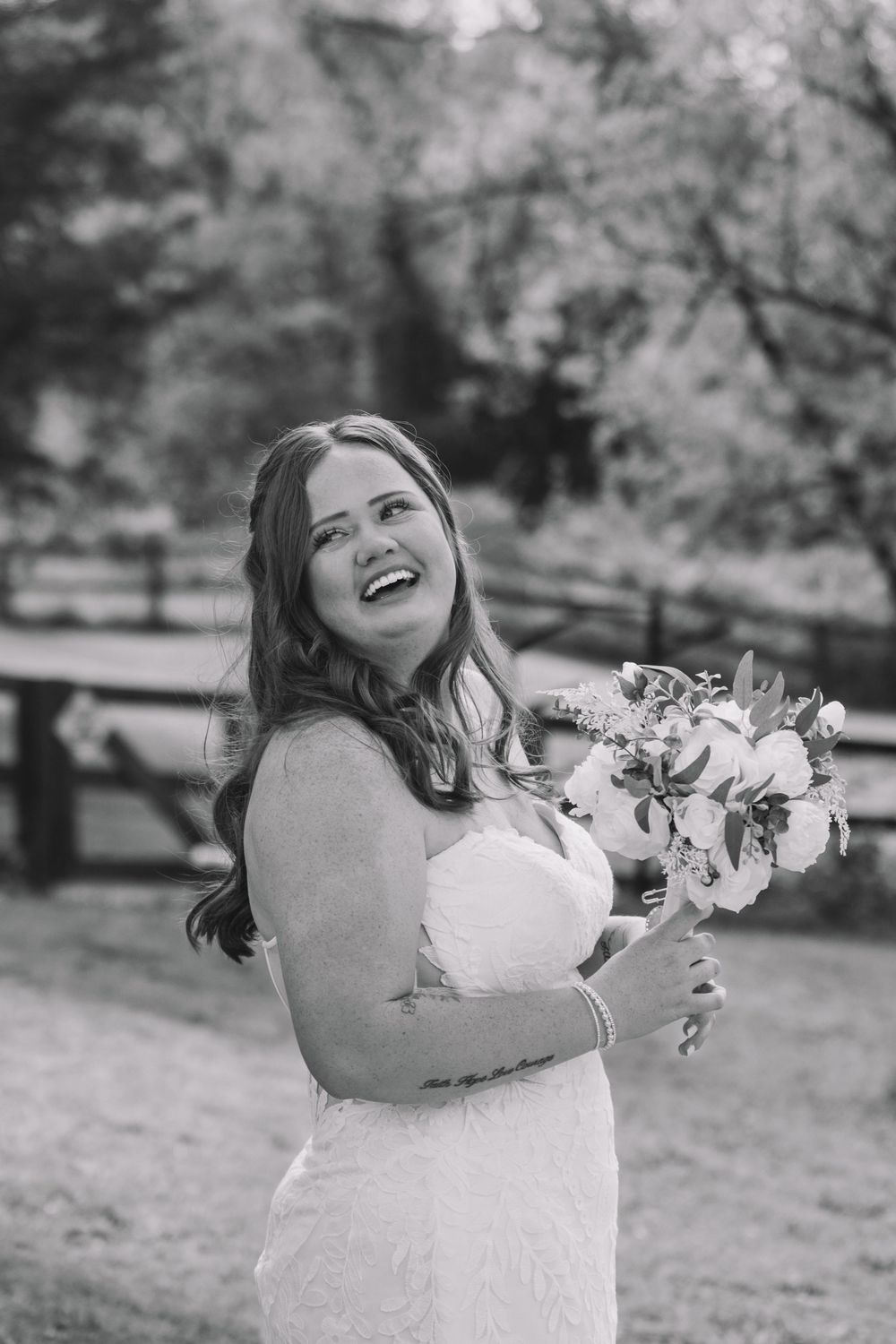 Black and white photo of a laughing bride in a strapless lace gown holding a bouquet outdoors.