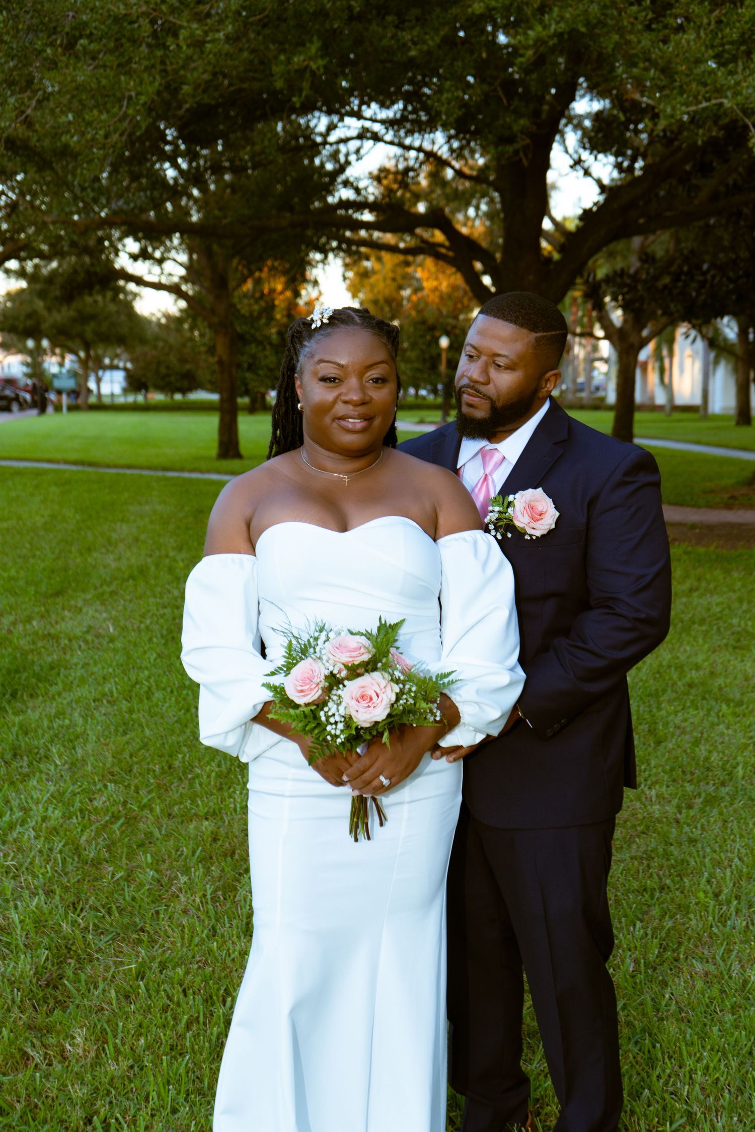 Newlyweds pose together under trees with bride holding pink floral bouquet.