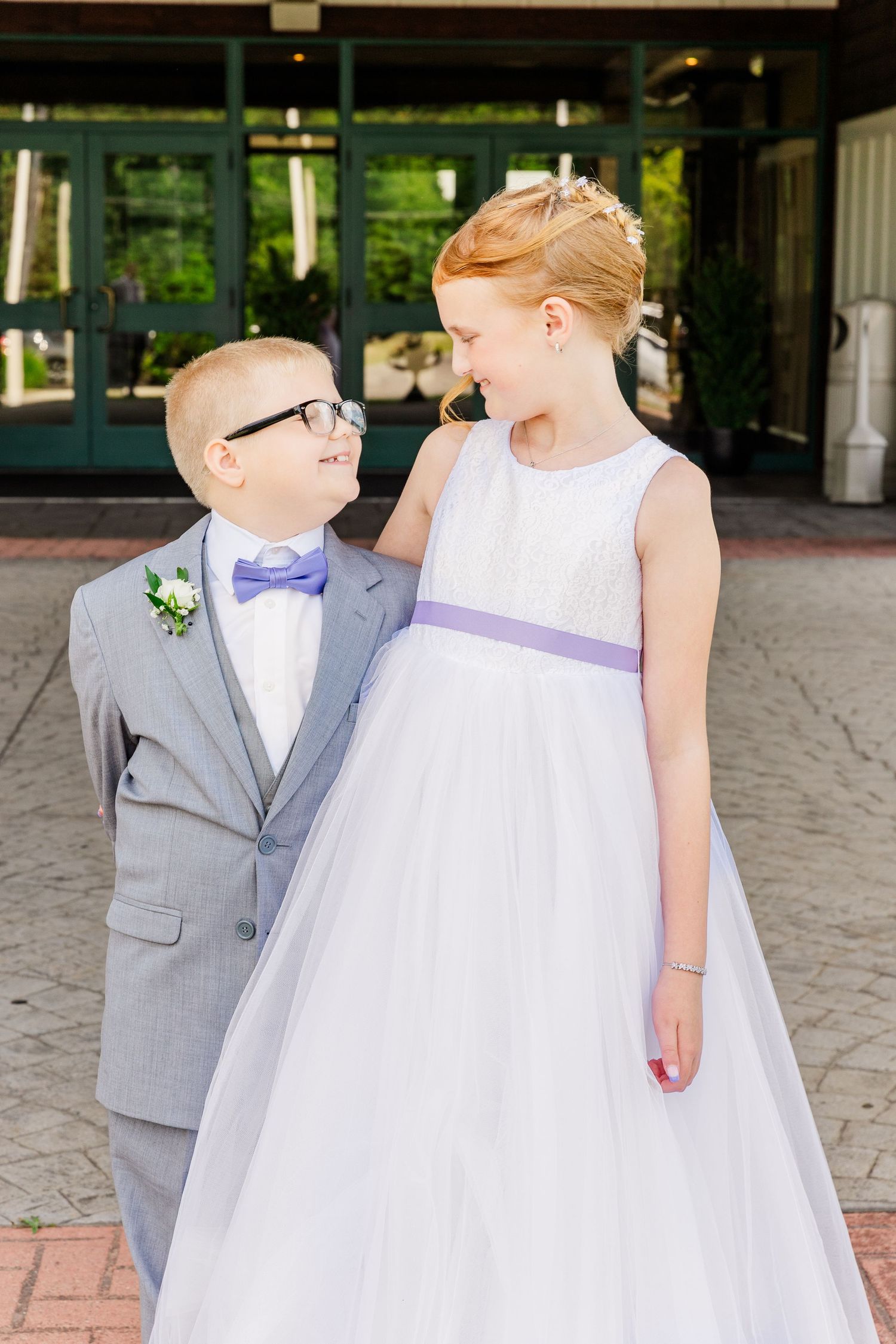 Heartwarming wedding moment between young attendants in gray suit and white dress with purple sash.