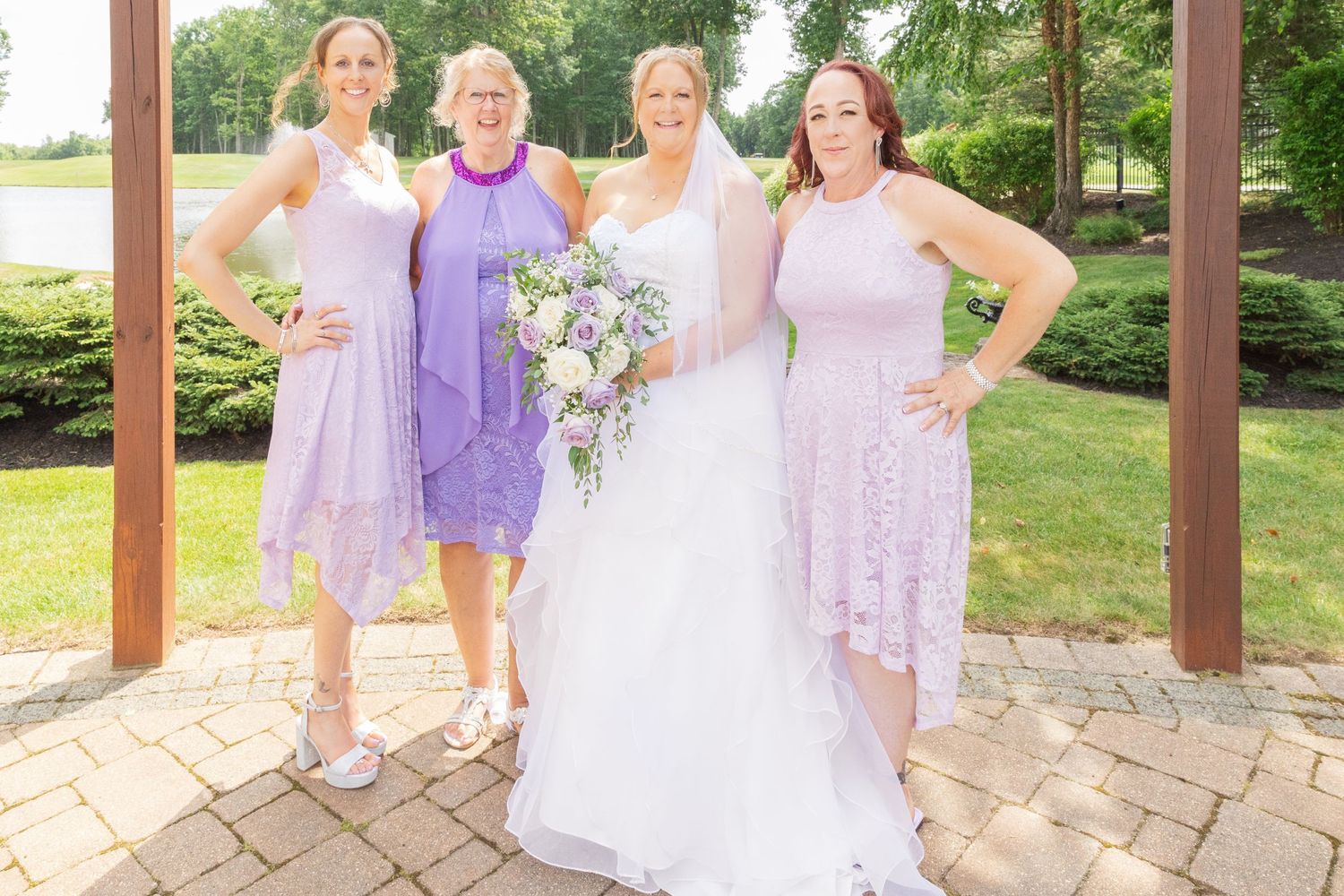 Wedding party in lavender dresses posing under wooden pergola in garden setting.