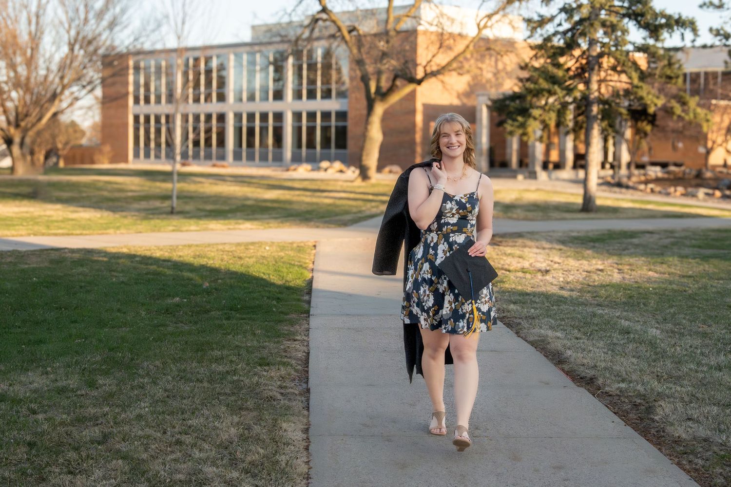 A girl holding a cap and gown walking down a sidewalk, smiling.
