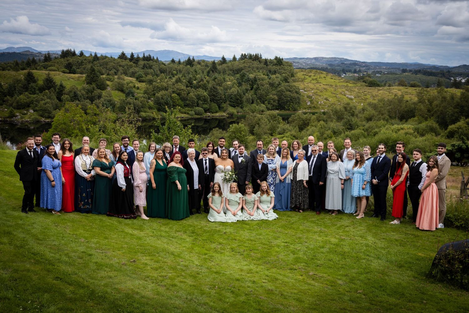 Large wedding party group photo on a lush green lawn with scenic countryside backdrop.
