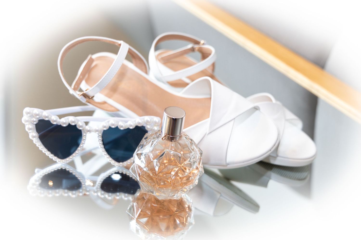 Wedding accessories flat lay featuring white heels, heart-shaped sunglasses, and perfume bottle.