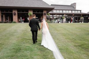 Newlyweds walking across a lawn towards a rustic venue building with guests in the background.
