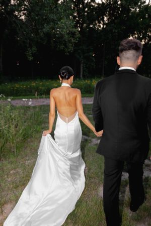 A couple walking through a garden path with the bride in a backless white gown and groom in a black suit.