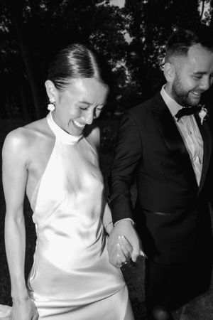Black and white photo of newlyweds sharing a joyful moment while holding hands outdoors.