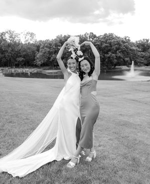 Two women in formal dresses celebrate outdoors, one in a bridal gown holding flowers aloft near a fountain.