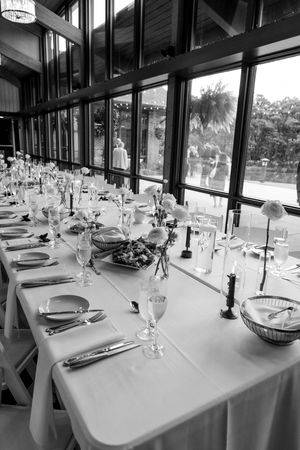 Black and white photo of an elegant wedding reception table set with fine china, glassware, and floral centerpieces by large windows.