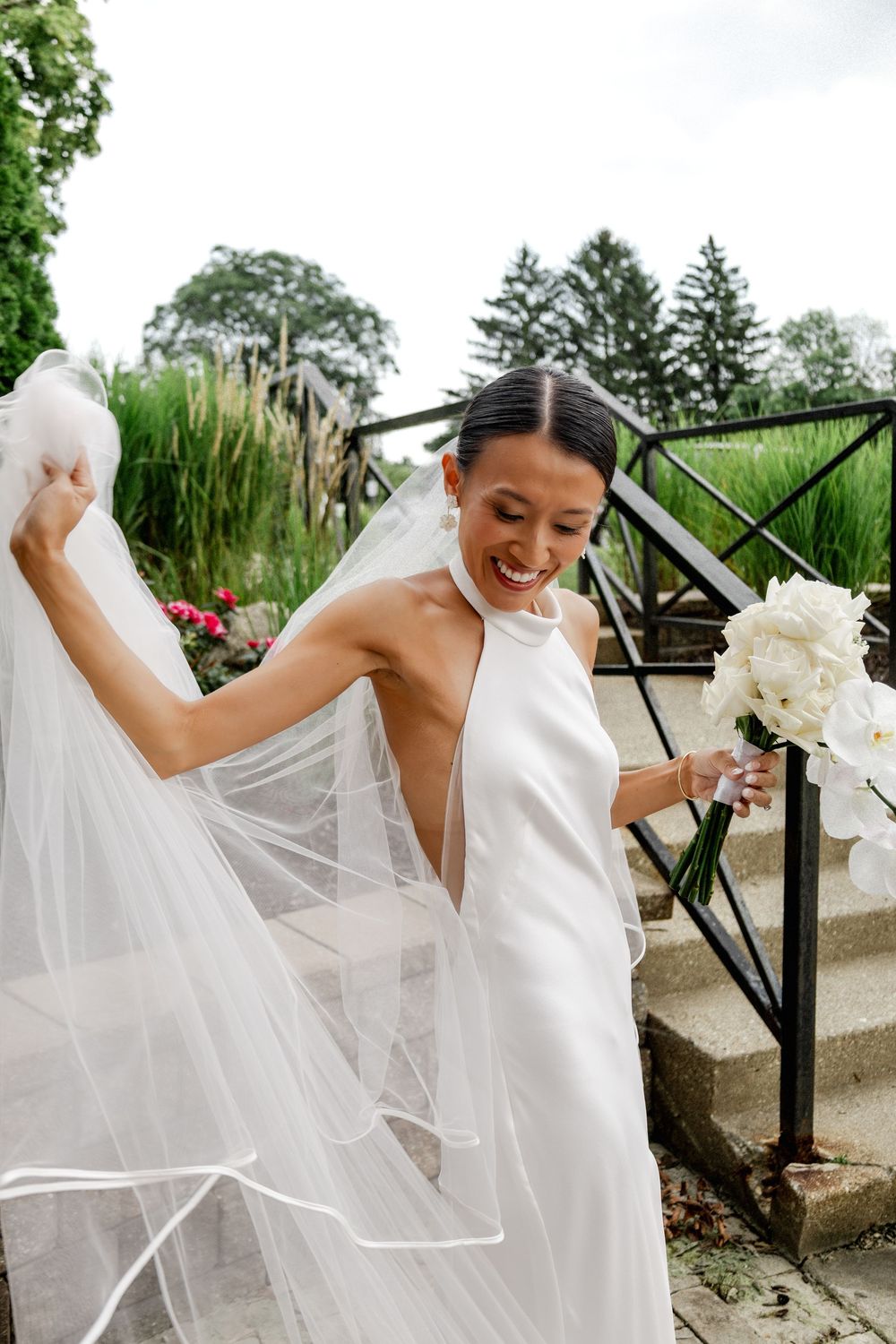 Joyful bride in white halter gown holding veil and bouquet on garden steps.
