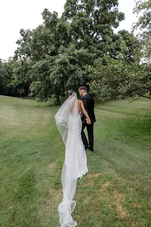 Bride and groom walking hand in hand on lush green lawn, bride's long white veil flowing behind her.