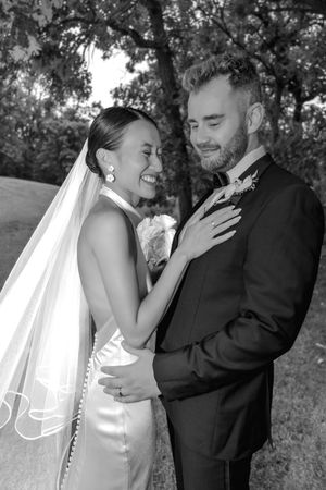 Black and white wedding photo of smiling bride in veil and groom in suit embracing outdoors among trees.