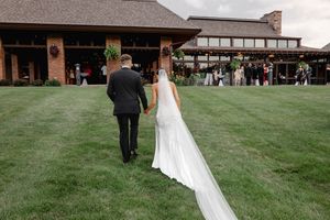 Bride and groom walk hand in hand toward elegant wedding venue on lush green lawn.