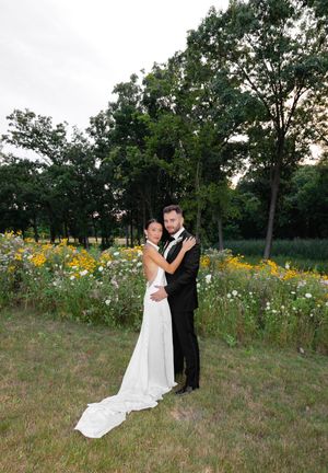 Bride and groom embracing in front of wildflower meadow and trees at golden hour outdoor wedding.