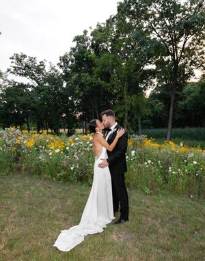Bride and groom share a romantic kiss in a lush garden with wildflowers and trees at sunset.