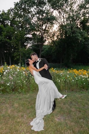 Groom lifts bride in white gown for a romantic kiss in a wildflower garden at sunset.
