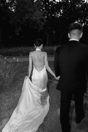 Black and white photo of bride in backless halter gown holding groom's hand walking toward trees.