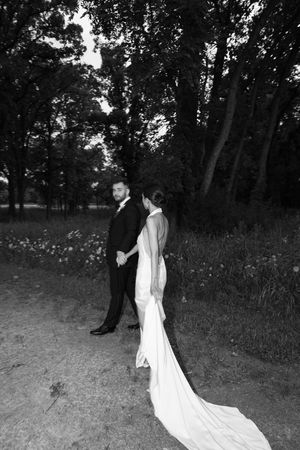 Black and white photo of bride and groom holding hands on a forest path, bride in elegant gown with long train.