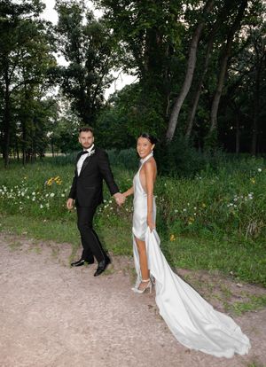 Bride and groom holding hands on a garden path, she in a white satin gown, he in a black tuxedo.