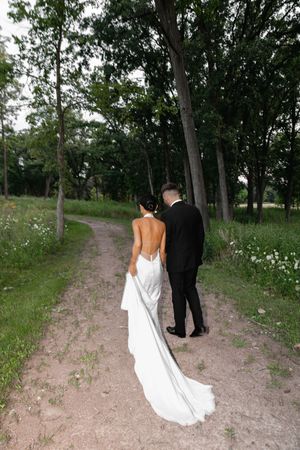 Bride and groom walking hand in hand down a dirt path surrounded by lush green trees in an outdoor wedding setting.