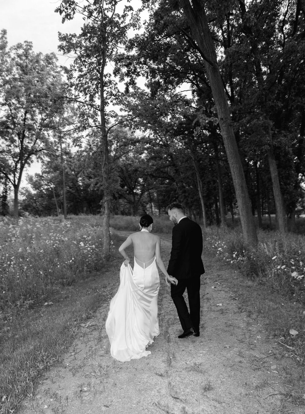 Black and white photo of bride and groom walking hand in hand along a tree-lined path on their wedding day.