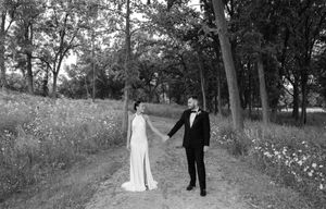 Black and white photo of a bride and groom holding hands on a tree-lined path in a romantic outdoor wedding setting.