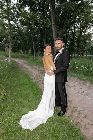 Bride in backless white gown and groom in black tuxedo pose together on a wooded path surrounded by lush greenery.